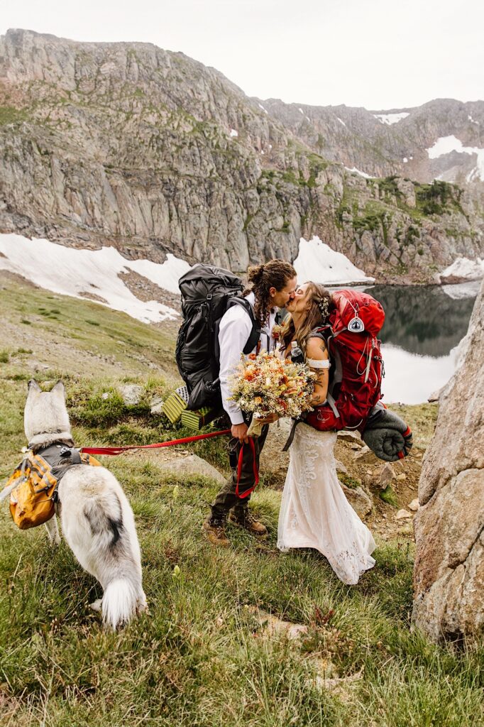 Bride and Groom kiss while hiking through the mountains with hiking backpacks and their husky during their Colorado Elopement Ceremony. 