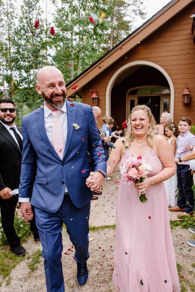 Bride in a beautiful pink dress walks down the aisle with her groom in a blue suit as their guests toss flower petals at them after their intimate wedding ceremony in Breckenridge Colorado. 