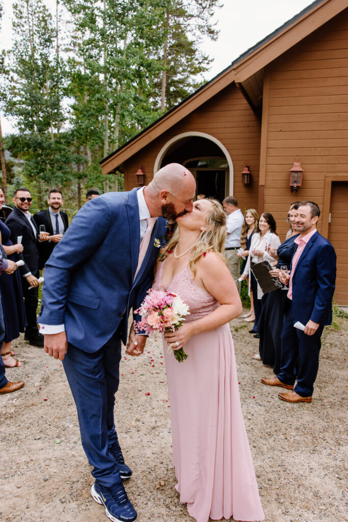 Bride in a beautiful pink dress kisses her groom in a blue suit as their guests toss flower petals at them after their intimate wedding ceremony in Breckenridge Colorado. 