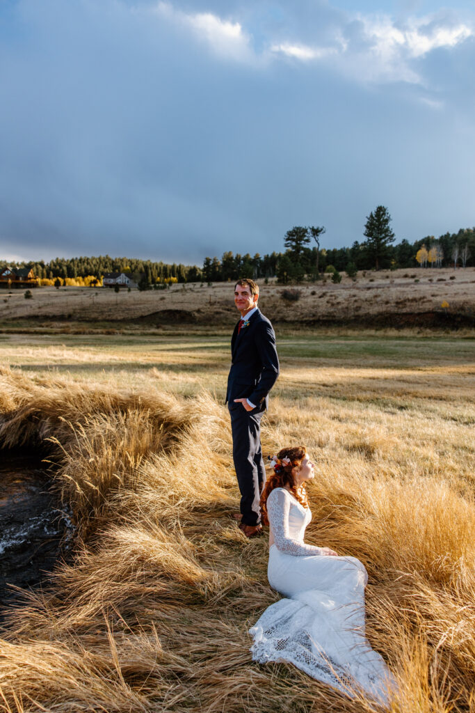 Bride with a flower crown lays in grassy plains as her groom stands over her for wedding portraits in Breckenridge Colorado. 