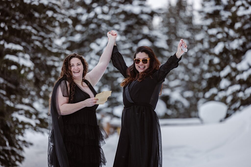Brides in all black gowns raise their hands in excitement posing amongst snowy trees after reading their vows at their Colorado Elopement  Ceremony. 