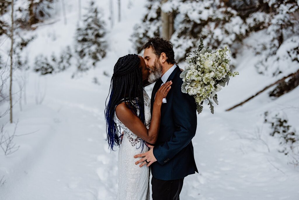 Bride holding an all white and green bouquet and Groom kiss with a snowy background during their elopement ceremony in Colorado. 