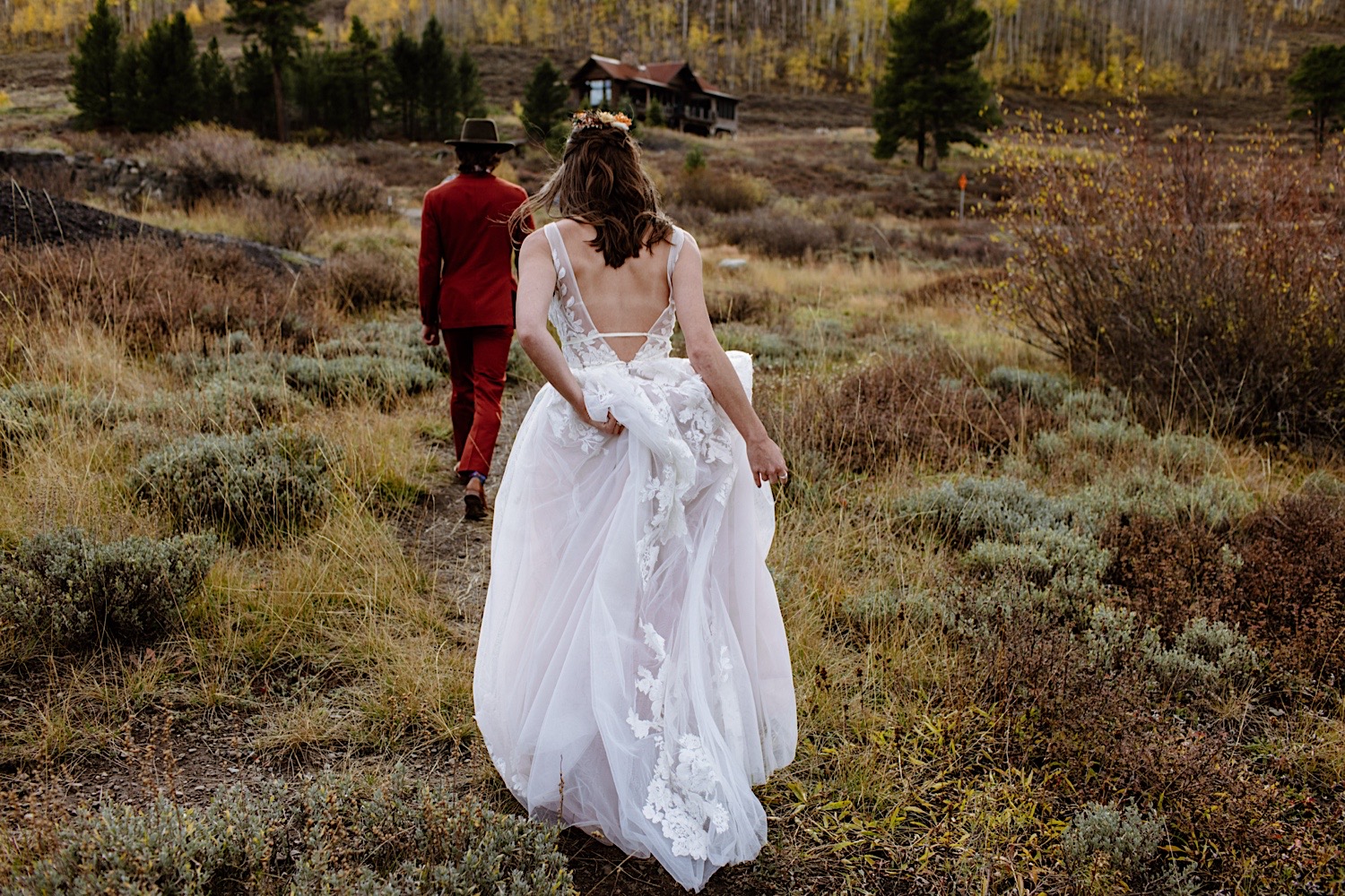 Bride in beautiful boho gown runs after her groom in an all red suit as they walk towards a lodge in Colorado for their elopement ceremony.