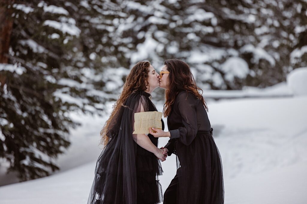 Brides in all black kiss with a snowy background during their elopement ceremony in Colorado. 