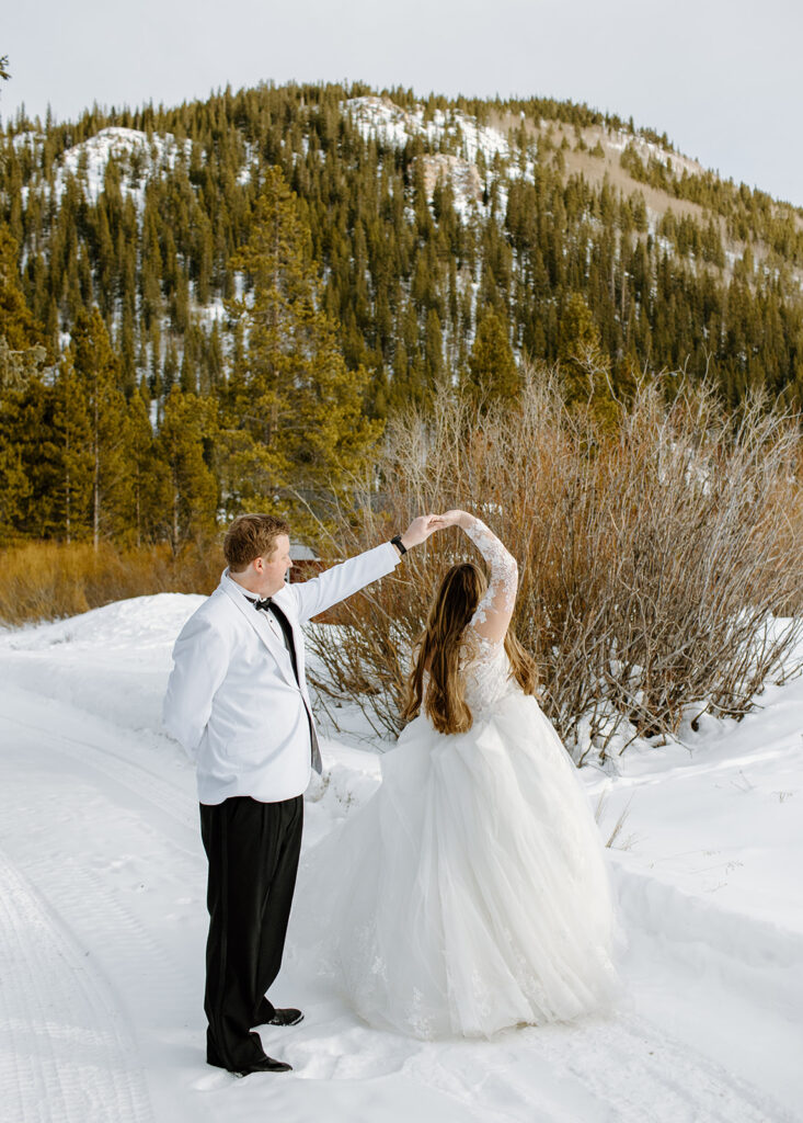 Bride in a lacy A-line dress and Groom in a white suit jacket and black pants dance in the snow with a mountain backdrop for wedding portraits in Breckenridge, Colorado. 