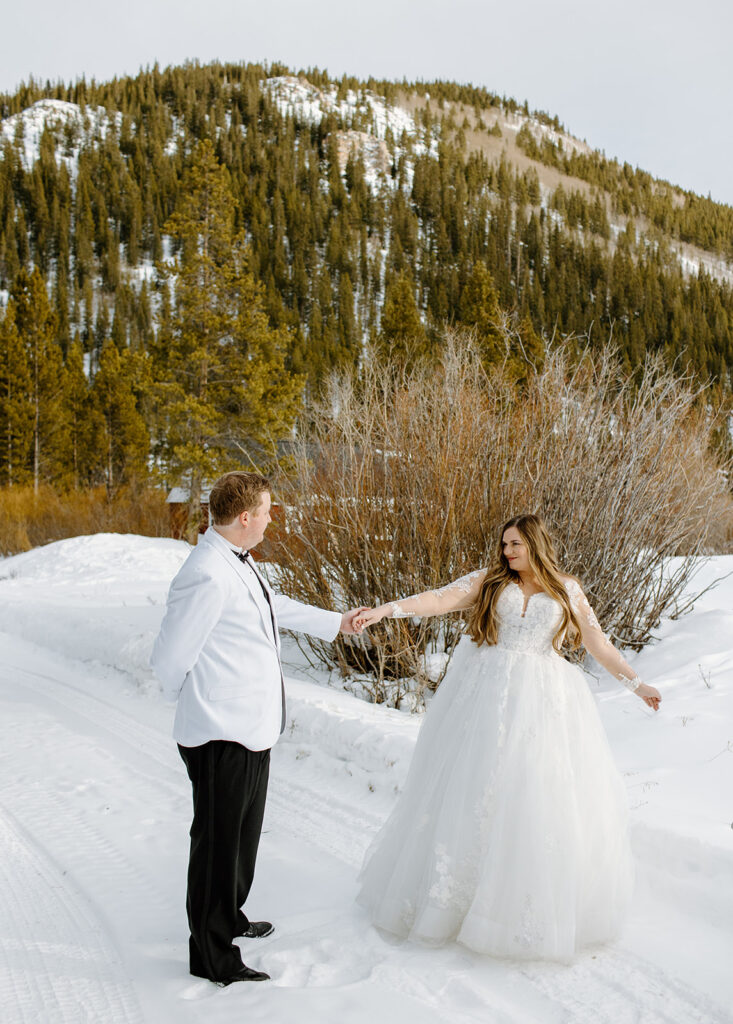 Bride in a lacy A-line dress and Groom in a white suit jacket and black pants dance in the snow with a mountain backdrop for wedding portraits in Breckenridge, Colorado. 